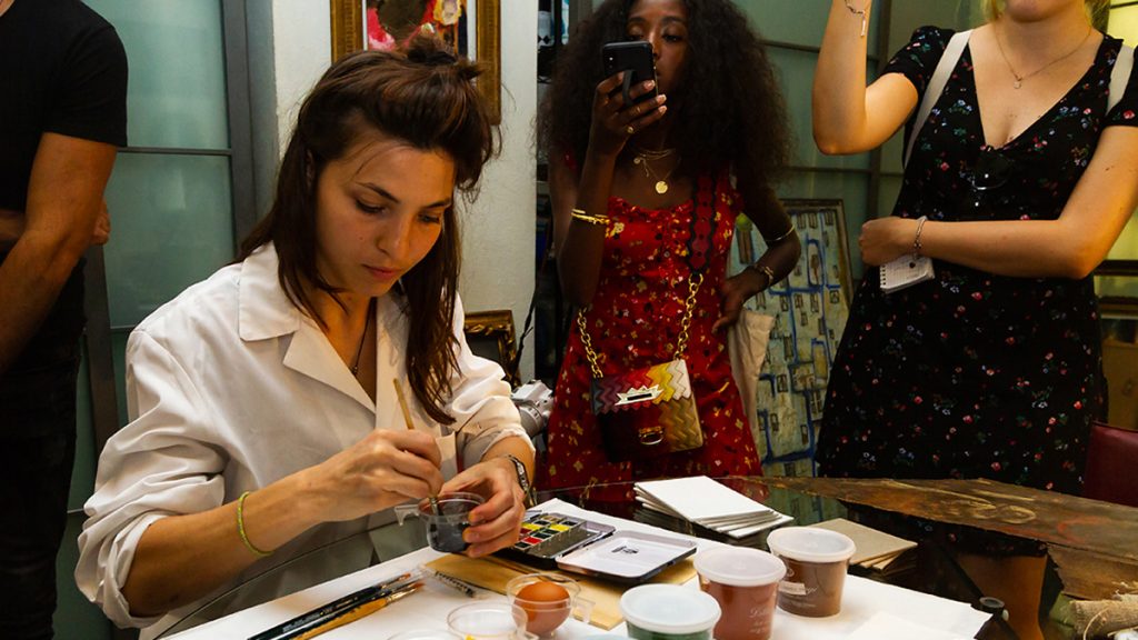 Young woman preparing colors for painting while other women observe and take pictures