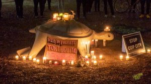 A stone turtle sculpture with candles and "Rise in protest Tortuguita" sign, and a "stop cop city" sign