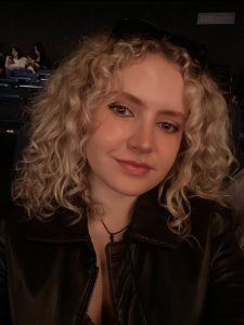 A white, femme presenting person with curly blonde hair wearing a black leather jacket